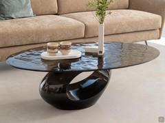 Table basse elliptique Dubai avec base sculpturale en finition laqué et plateau en verre transparent, conçue pour l'avant d'un canapé.